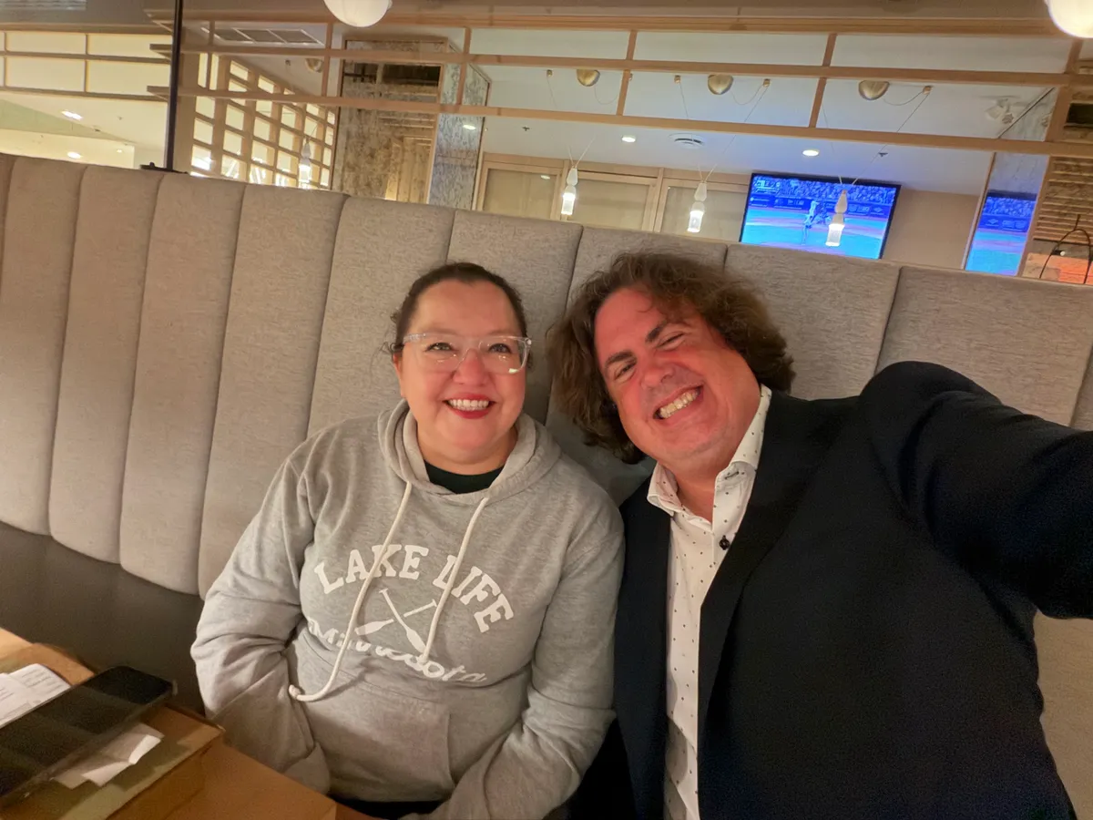 National Chief Cindy Woodhouse Nepinak, in a grey "Lake Life" hoodie and clear glasses, and Philippe Beaudoin, in a dark blazer, laughing side by side on a grey restaurant booth in Ottawa.