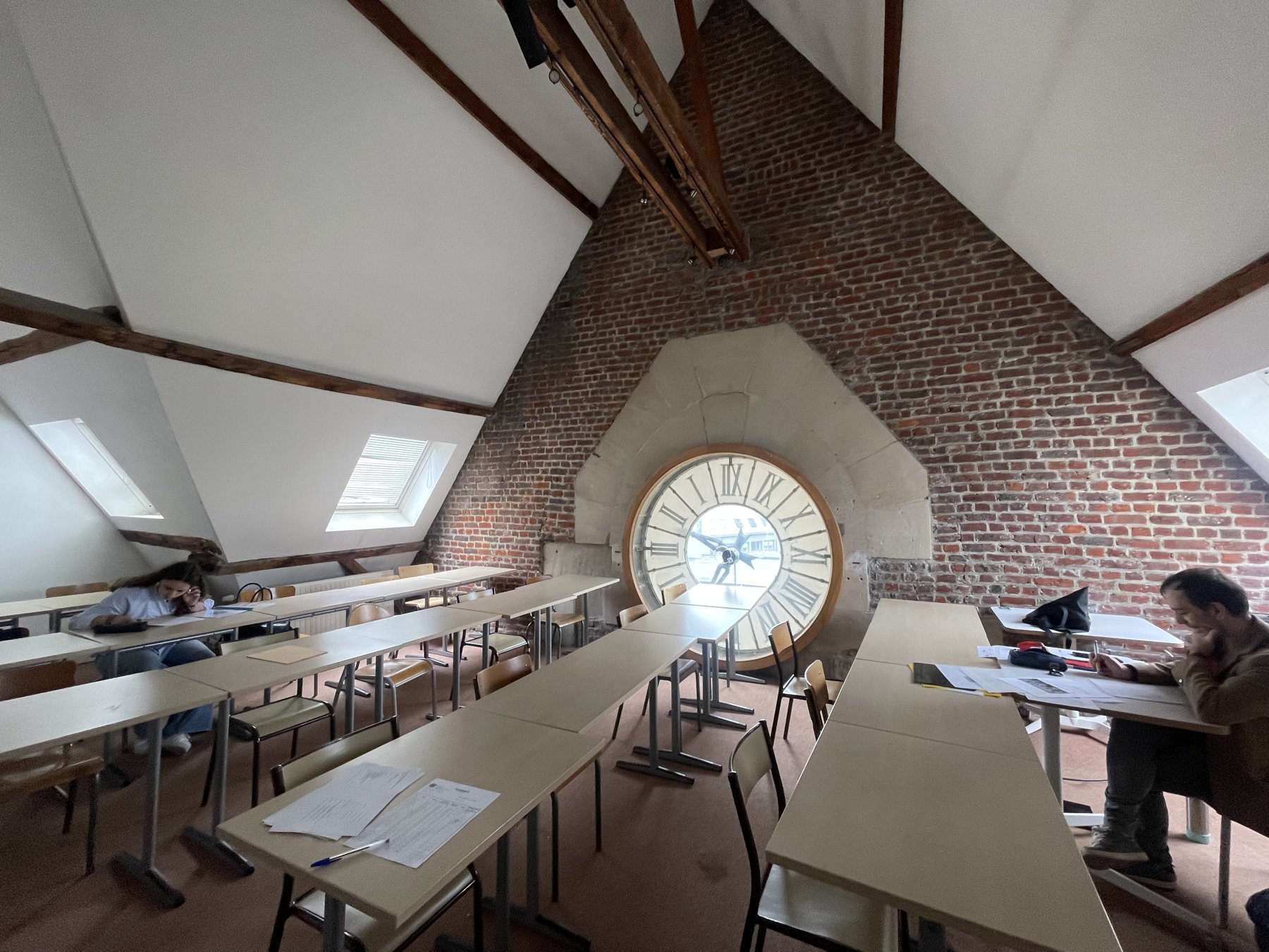 Une salle de cours sous les combles : poutres de bois, pignon de briques rouges, deux lucarnes, des tables claires en rangées, deux étudiants qui travaillent. Au fond, encastré dans le mur de briques, un grand cadran d'horloge blanc vu de l'intérieur, avec ses chiffres romains à l'envers.