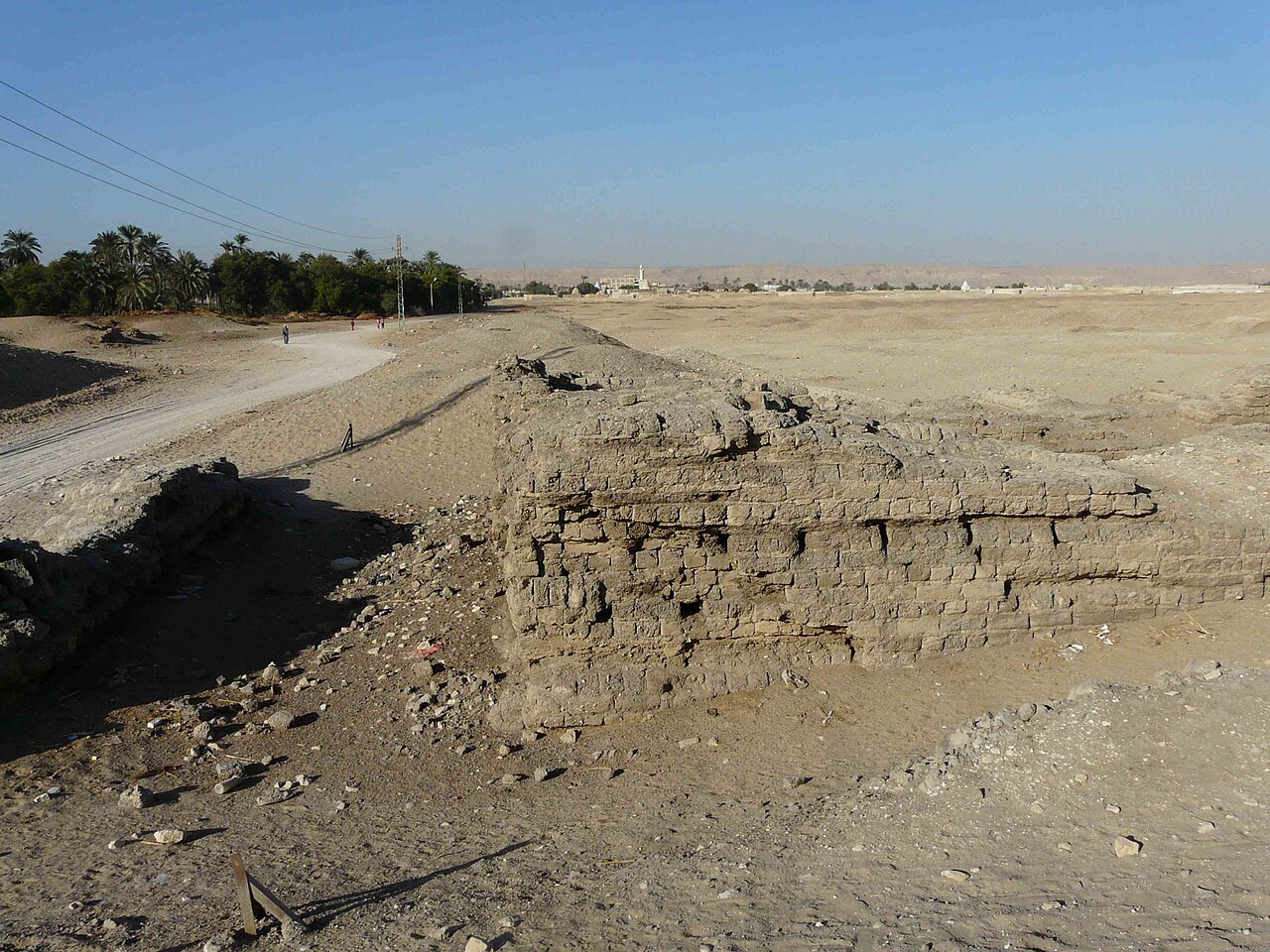 A thick stump of dark mud-brick wall, taller than a person, standing alone beside a pale dirt track under electricity wires; beyond it flat sand stretches to a distant village with a minaret and a line of cliffs.