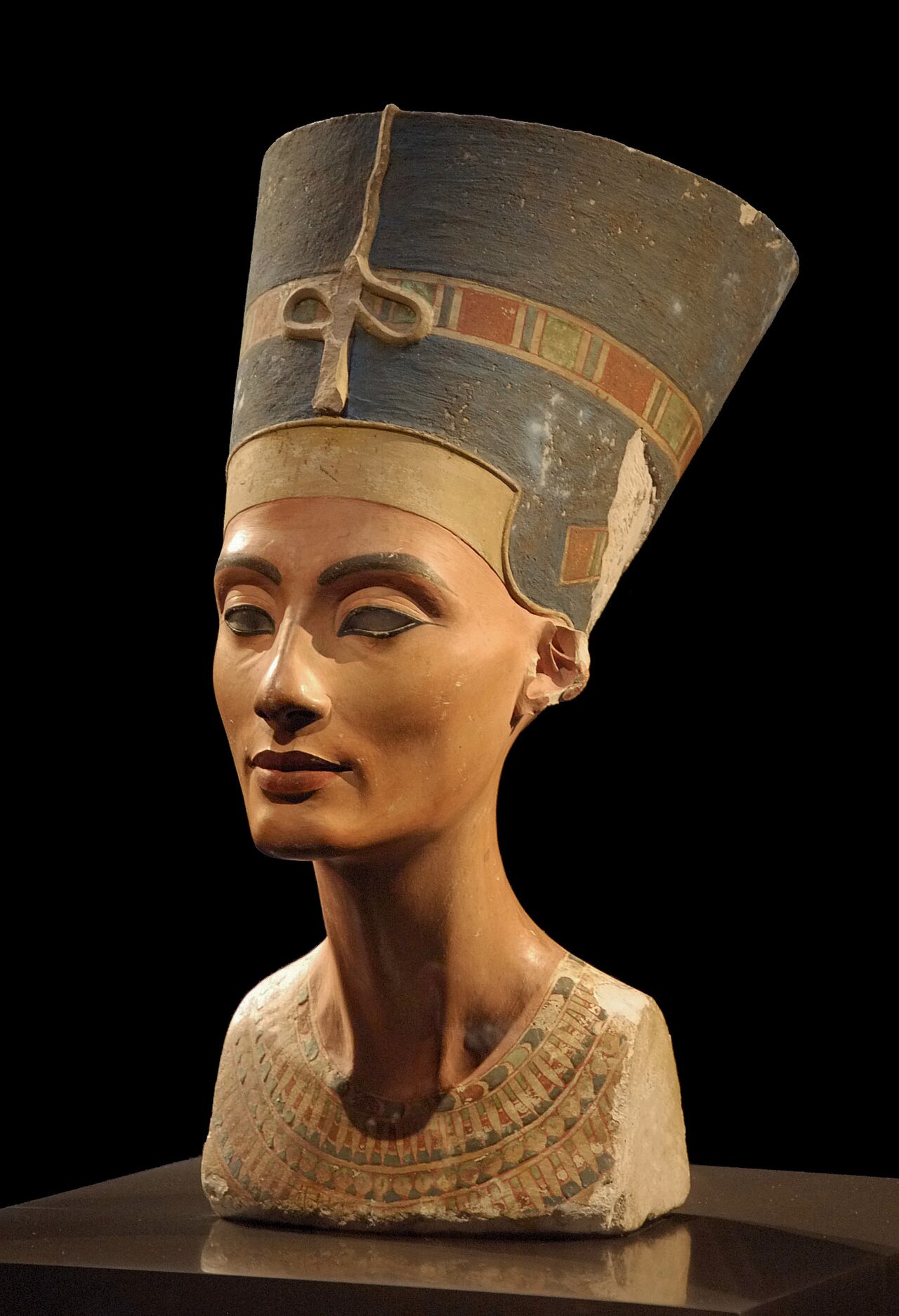 Against a black background, the painted limestone bust of Nefertiti: a long neck, a calm brown face with dark-lined eyes, one of them without an iris, and a tall flat-topped blue crown with a coloured band, lit from one side.