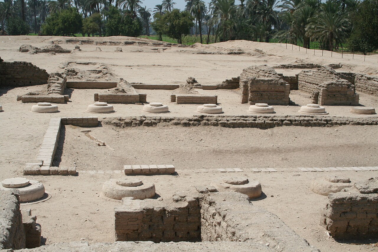 Low walls of dark mud brick laid out in rectangles on pale sand, with two rows of round white limestone column bases standing in the open, and behind them a dense line of palm trees and green fields.