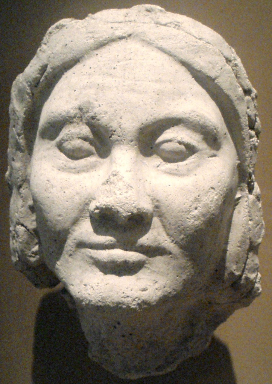 A life-sized plaster face of an old woman, pale and pitted, seen straight on: heavy lids, pouches under the eyes, deep lines from the nose to the corners of the mouth, a lined forehead, the mouth set. The hair is a rough band; the face is calm.