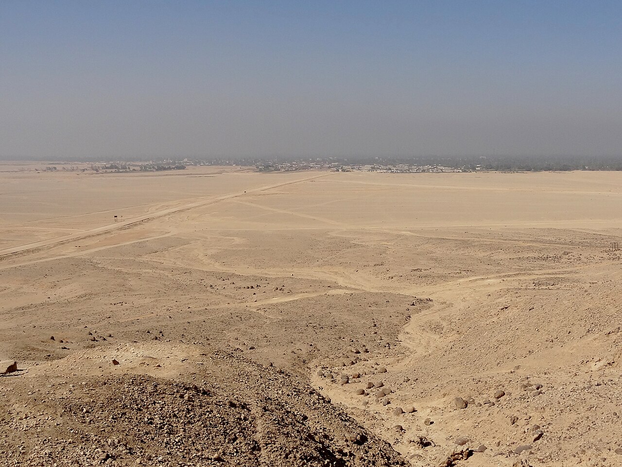 Seen from a rocky slope: a vast flat plain of pale sand under a hazy sky, crossed by two thin tracks that meet far off, and at the horizon a dark line of palm trees and low village buildings along the river.