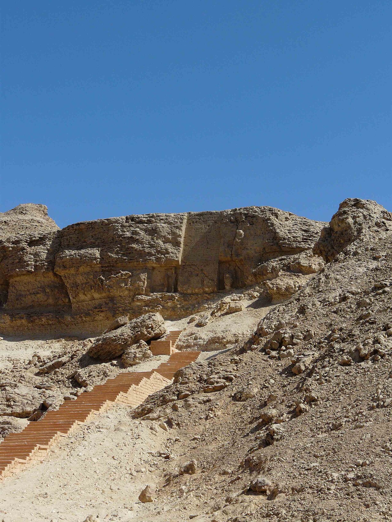 Under a deep blue sky, a pale rocky cliff with a tall smoothed rectangular panel cut into its face near the top, and a modern brick staircase climbing steeply up the scree toward it.
