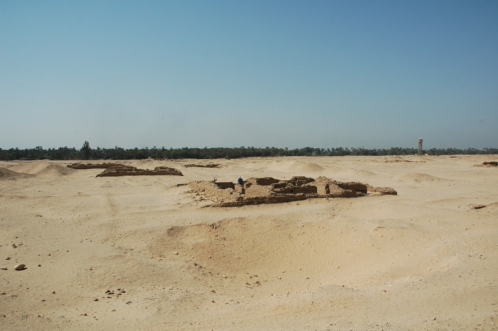 A wide expanse of pale sand under a clear sky, with a low cluster of eroded mud-brick walls in the middle distance and two small figures sitting among them; a line of palms and a lone water tower on the horizon.
