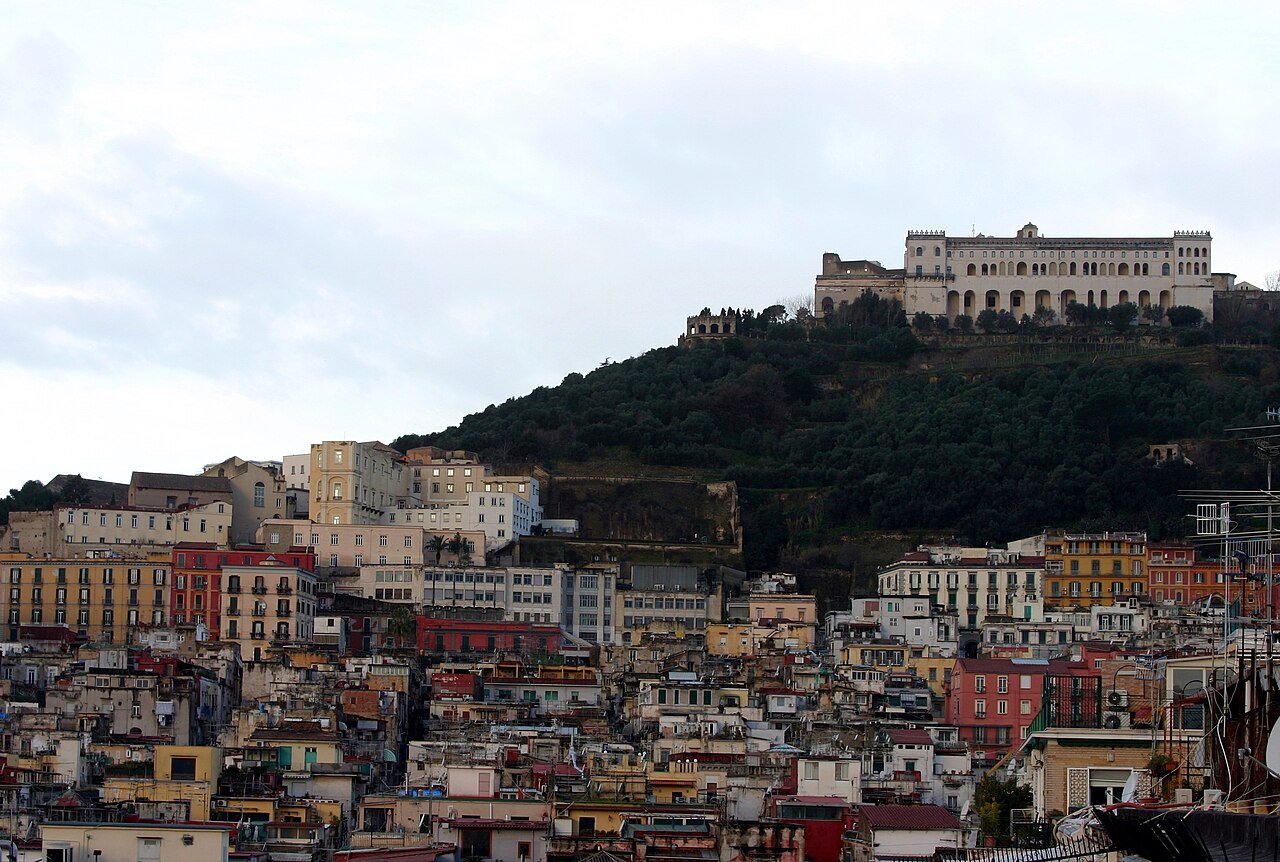The white mass of the Certosa di San Martino on its green hill, above the dense stacked houses of the Quartieri Spagnoli, under a grey sky.