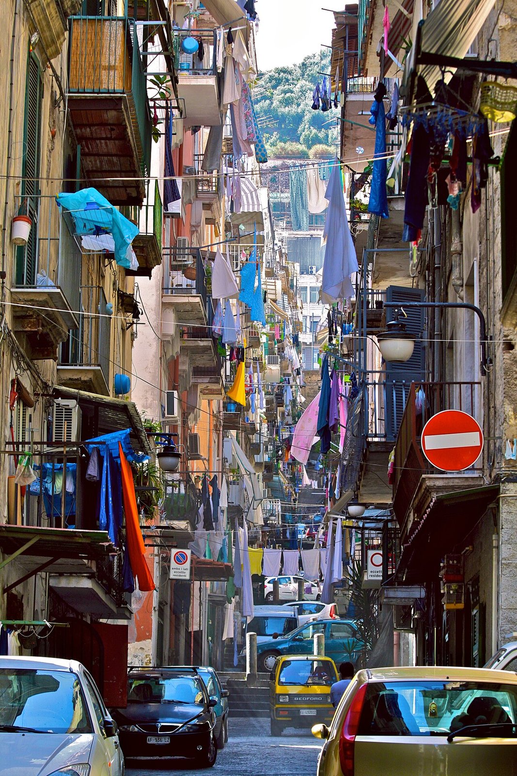 A steep lane of the Quartieri Spagnoli seen from above, lines of washing strung from balcony to balcony all the way down, cars parked below, a green hill closing the view at the top.