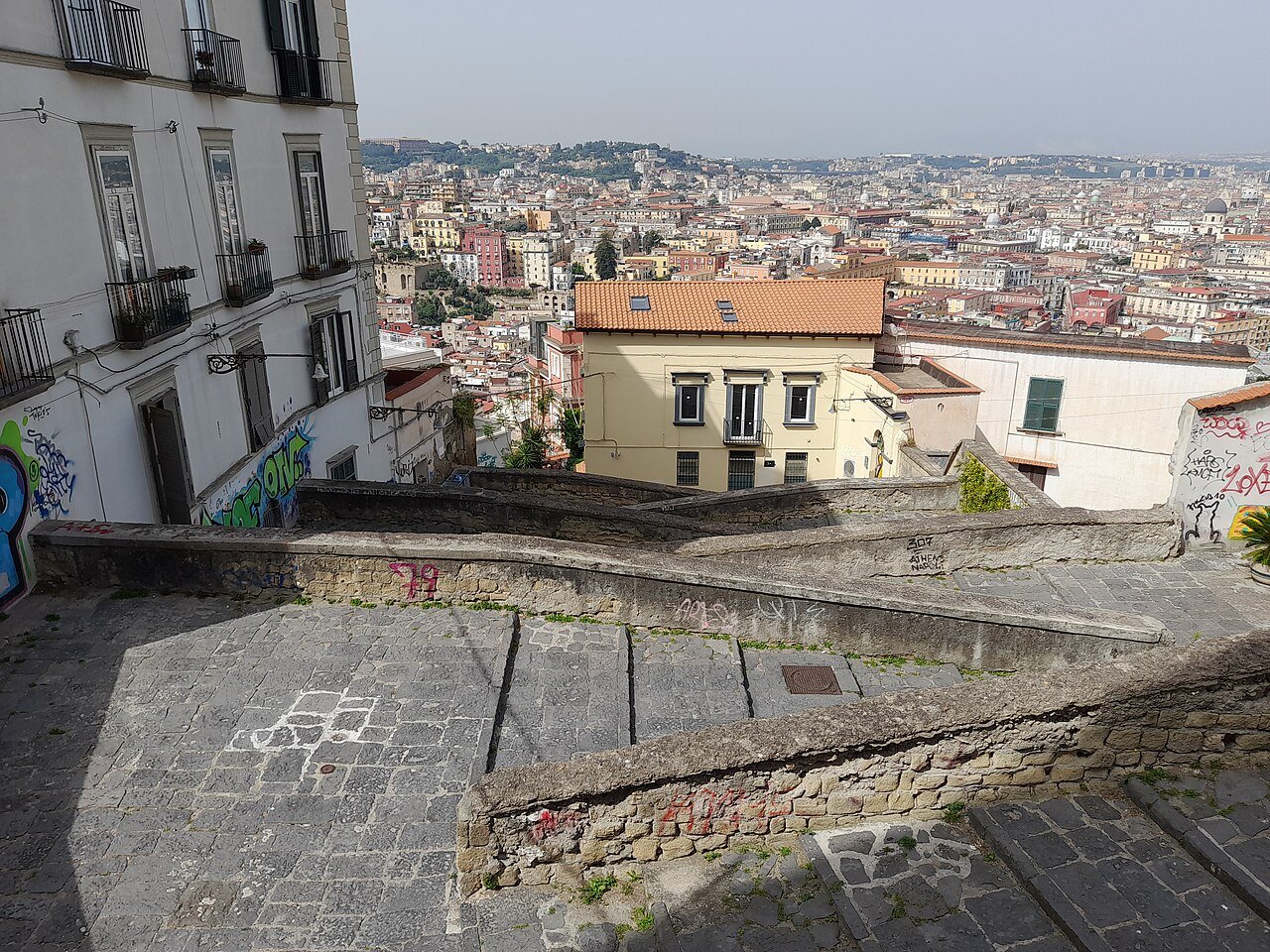 From a stone landing on the Pedamentina, the roofs of Naples spread below in every direction, terracotta and white, fading into haze toward the far hills.