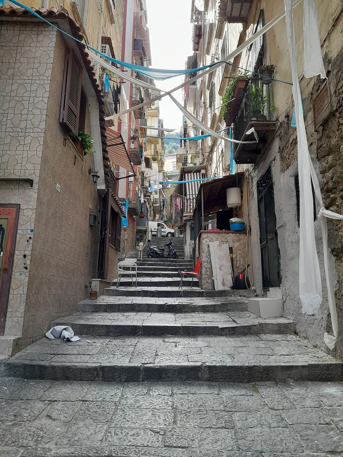 A stepped lane climbing between tall houses in the Quartieri Spagnoli, strings of small flags crossing overhead, a cloth hanging from a balcony, sky at the top.