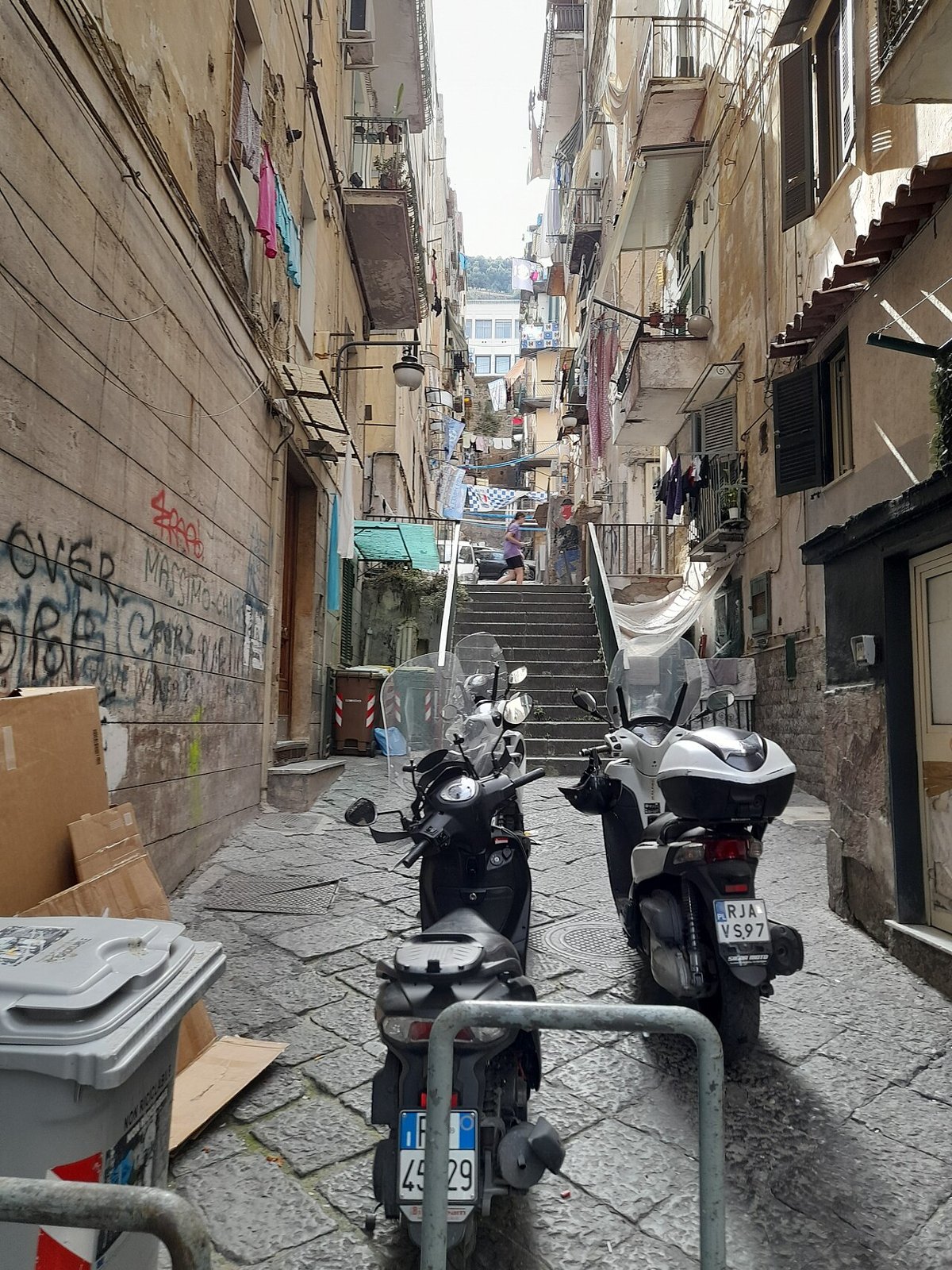 A narrow lane in the Quartieri Spagnoli climbing away between tall grey buildings, scooters parked along the wall, steps rising at the far end into light.
