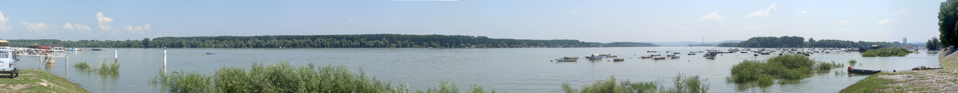 A very wide panorama taken from the Zemun quay: the flat grey-blue Danube filling the frame from left to right, a low green wooded shore on the far side, moored boats and barges near the near bank, and a pale hazy sky.