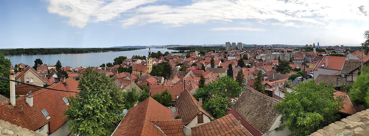 A wide view from the top of a hill: red tile roofs and a baroque church spire below, the broad grey Danube curving away to the left with a wooded island in it, and on the far bank, small under a big sky, the towers of Belgrade.