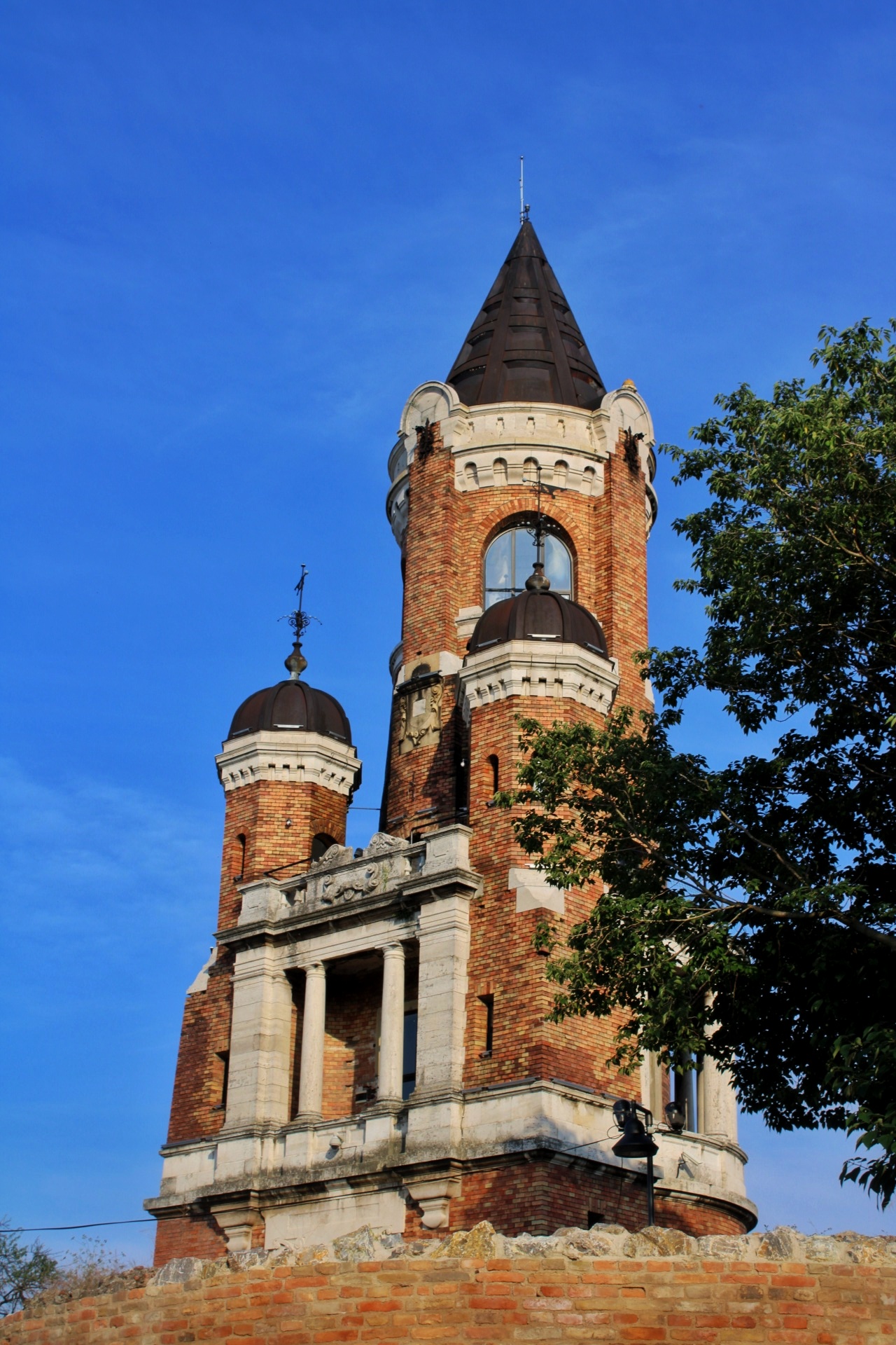 A tall slender tower of red brick and pale stone seen from below against a deep blue sky: a pointed dark cone on top, arched windows, four small turrets with dark onion caps at its base, standing on a low ruined wall of rough stone and brick.