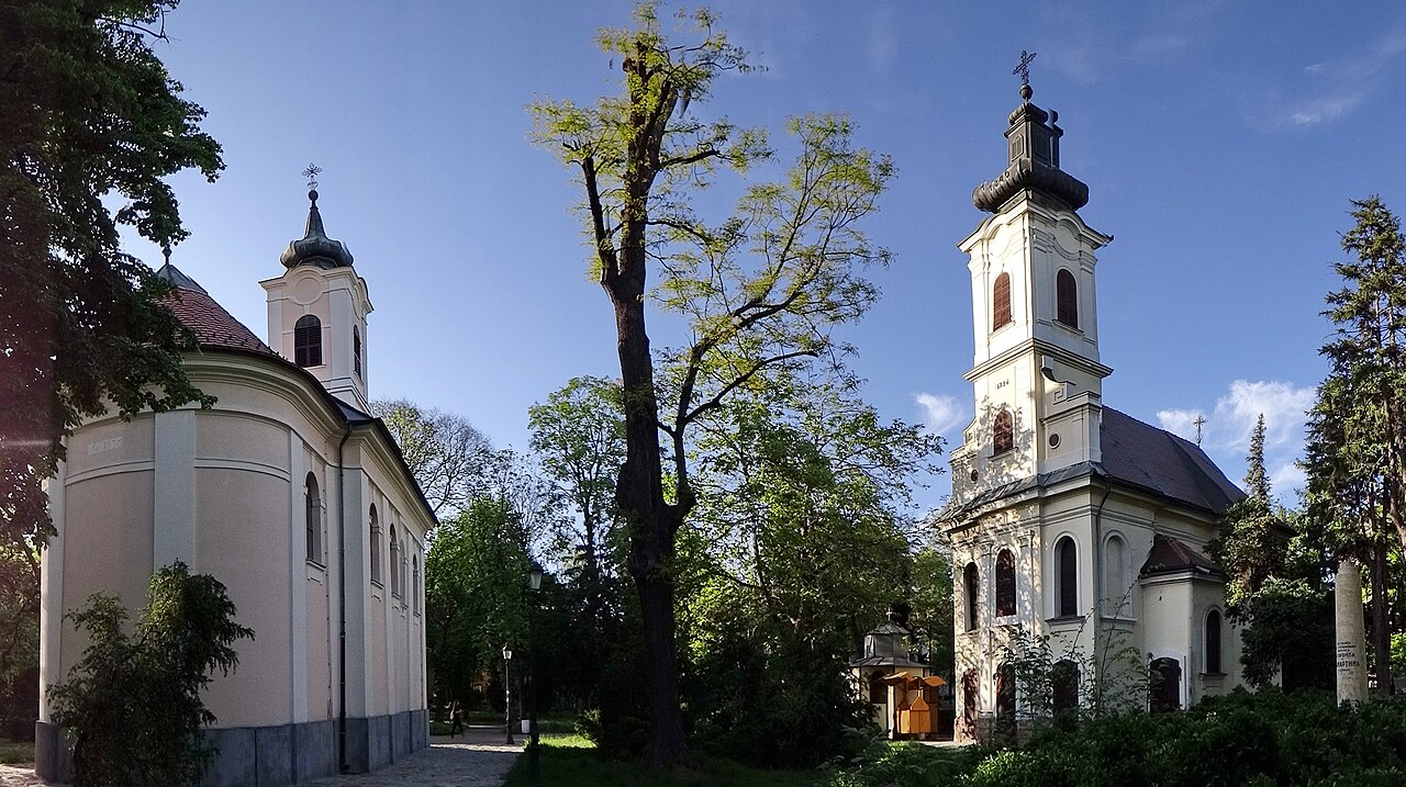 Two small baroque churches facing each other across a park path under old trees: on the left a cream-coloured chapel with a rounded apse and a little onion-topped bell tower, on the right a taller one with a black onion dome, blue sky above.