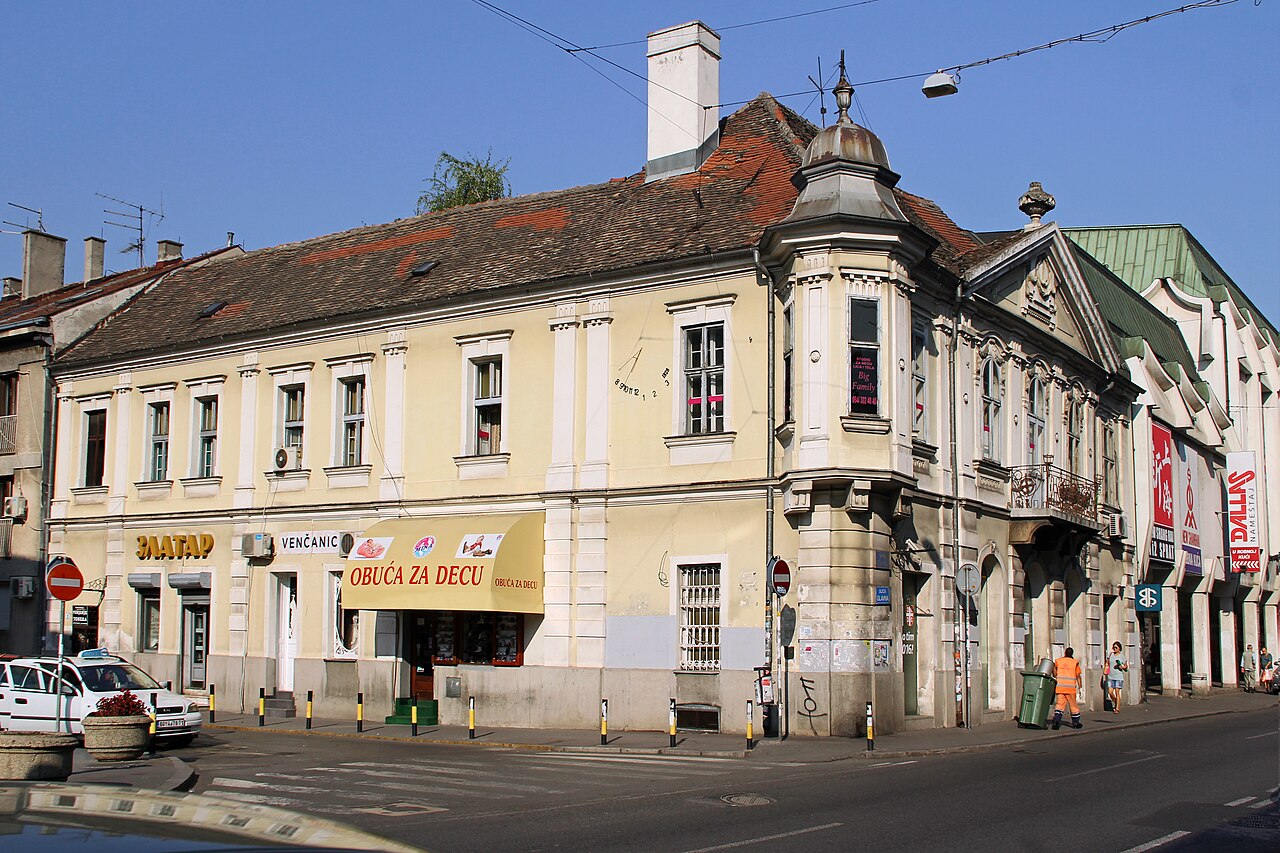 A yellow two-storey corner house with white pilasters and a small turret on the corner, shop awnings at street level, and on the upper wall between two windows the faint painted lines and numerals of a sundial.